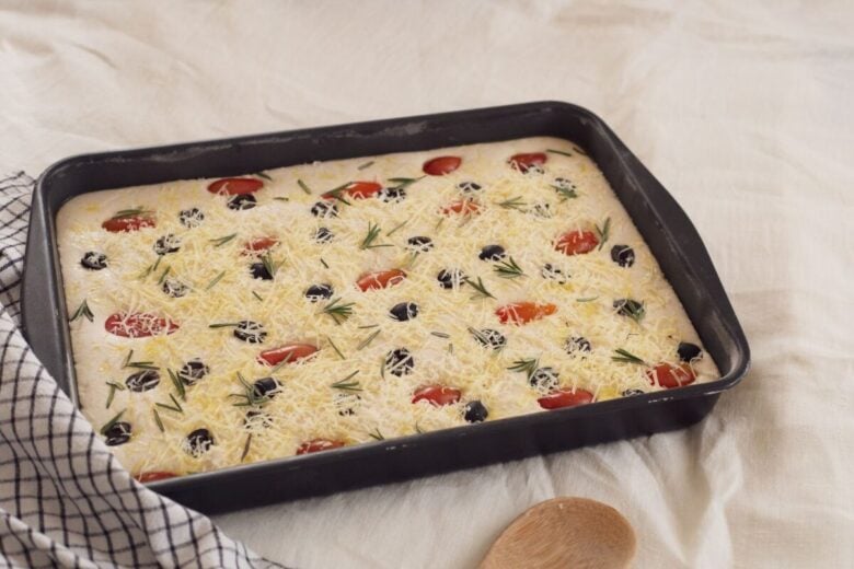 Unbaked focaccia dough topped with cheese, olives, tomatoes, and rosemary.