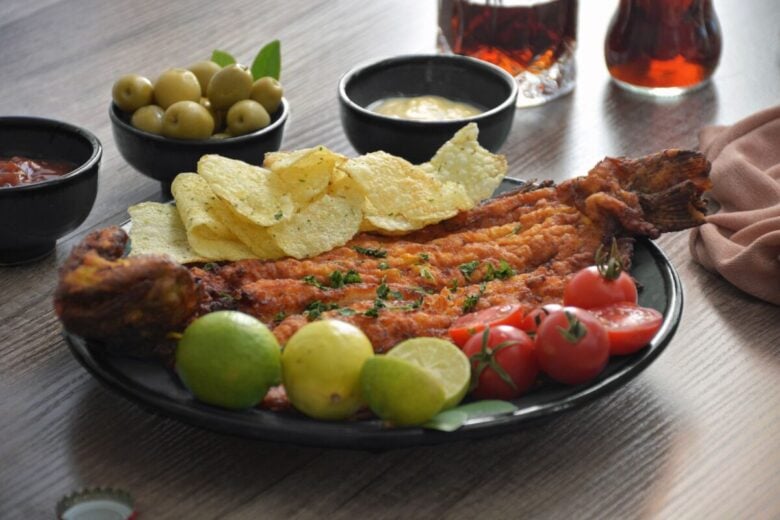 From above of tasty fried fish with crispy potato chips near fresh cherry tomatoes and limes on wooden table with green olives and sauces