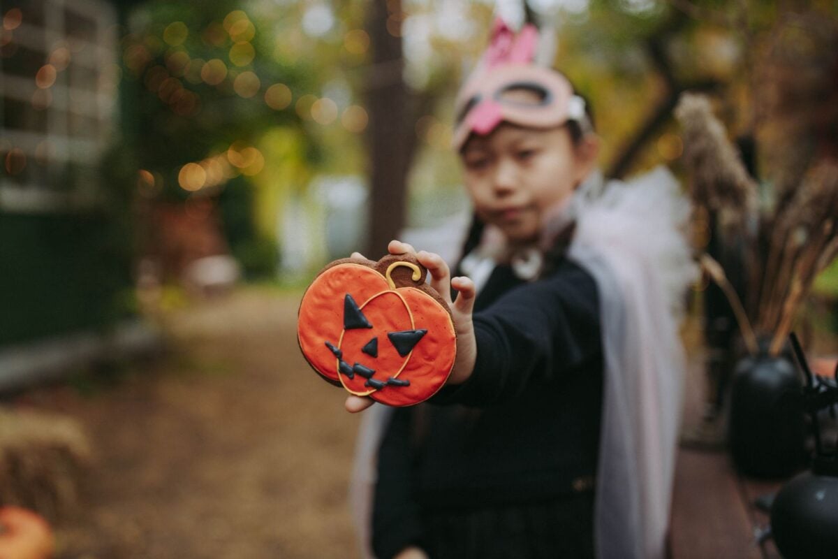 A child wearing a costume mask holds a Halloween-themed pumpkin cookie outdoors. A child wearing a costume mask holds a Halloween-themed pumpkin cookie outdoors.
