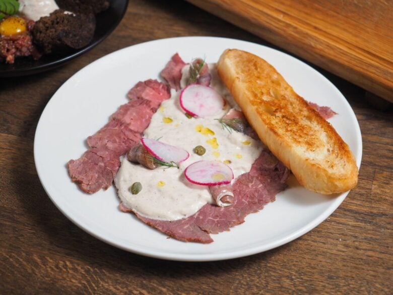 Sliced beef with creamy sauce and radishes on a white plate, served with toasted bread.